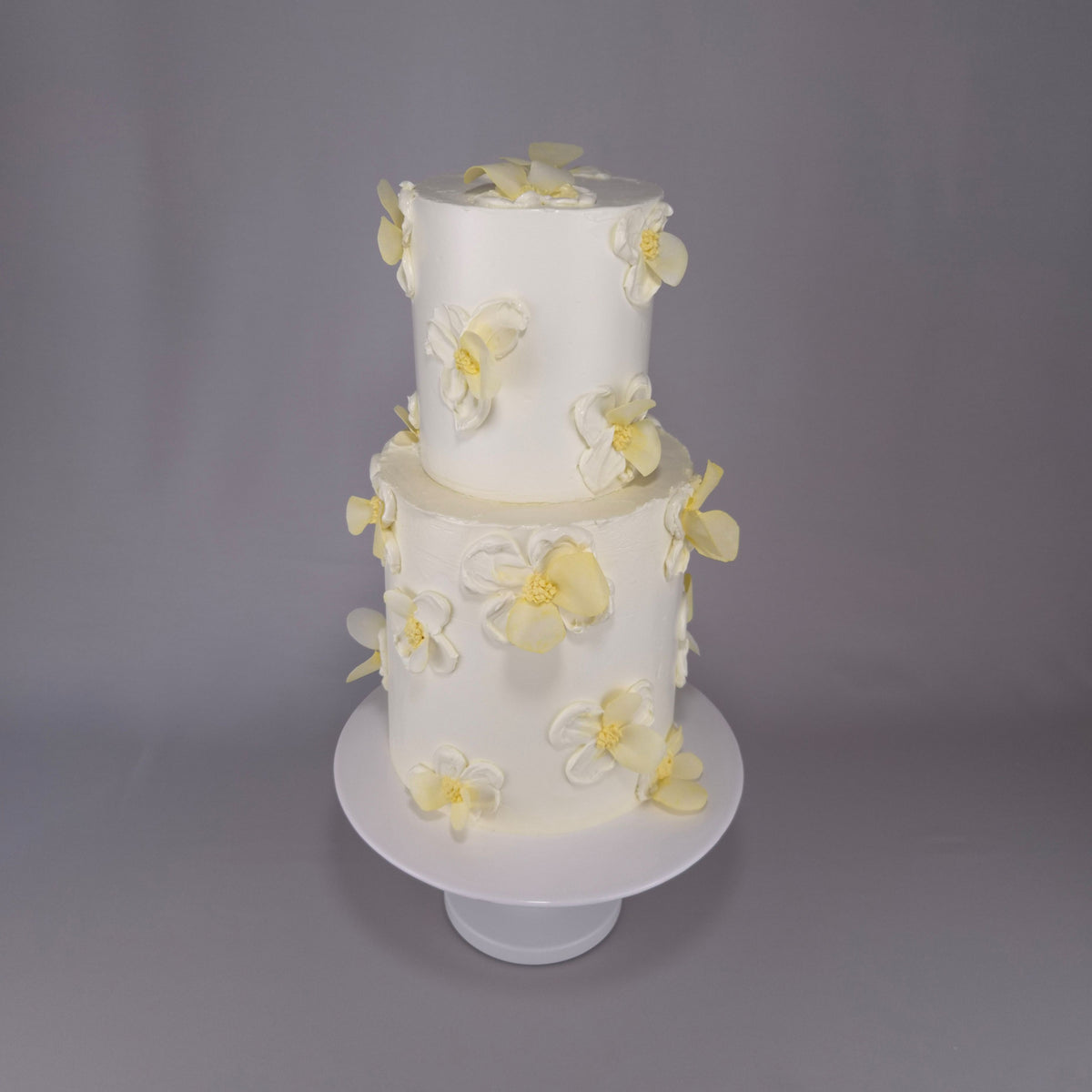 Zweistöckige weiße Tauftorte mit weißen und hellgelben Blüten auf einem weißen Tortenständer