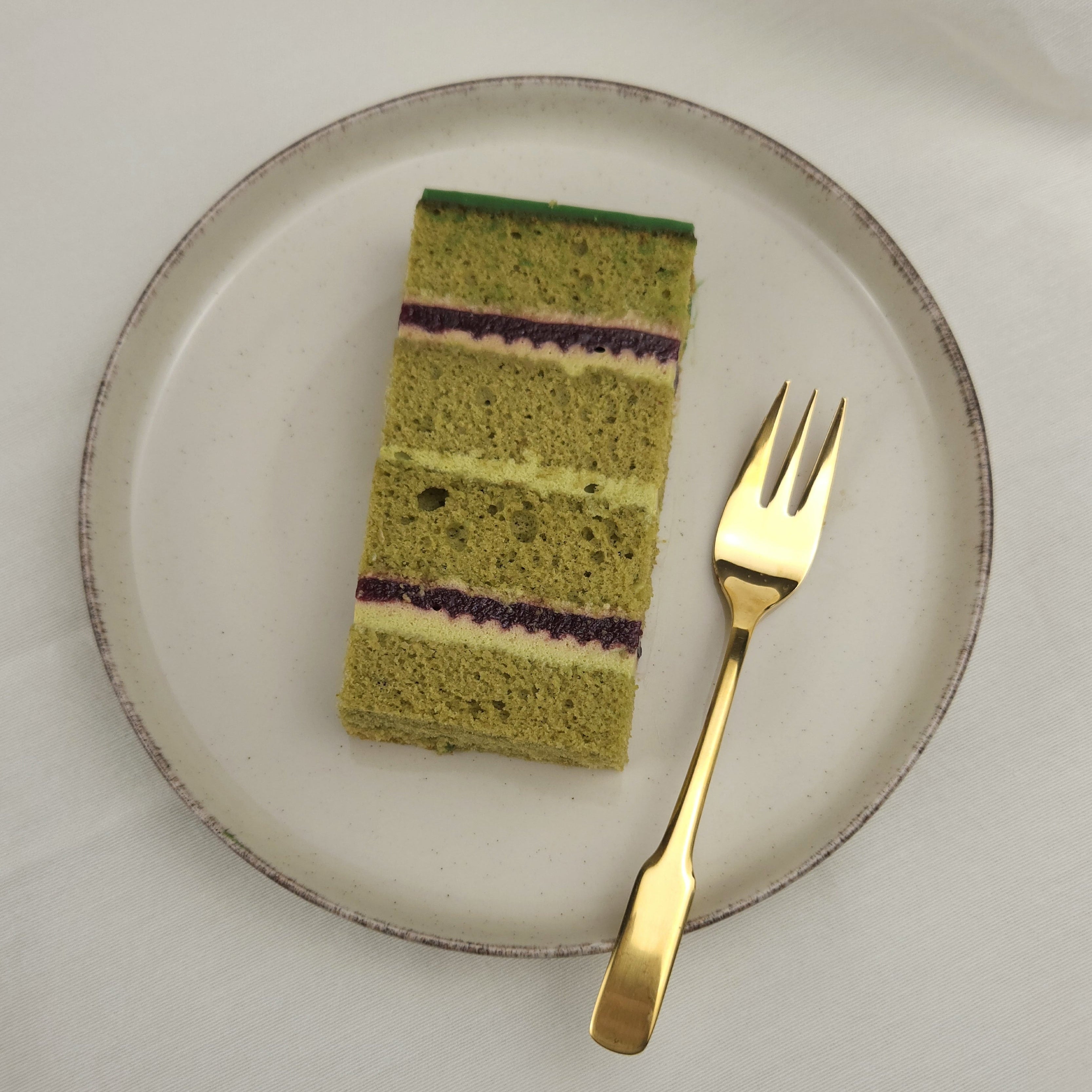 tück Matcha-Pistazien-Kirsch Torte auf Keramikteller mit goldener Kuchengabel – ein harmonisches Spiel aus grünem Tee, Nuss und Frucht