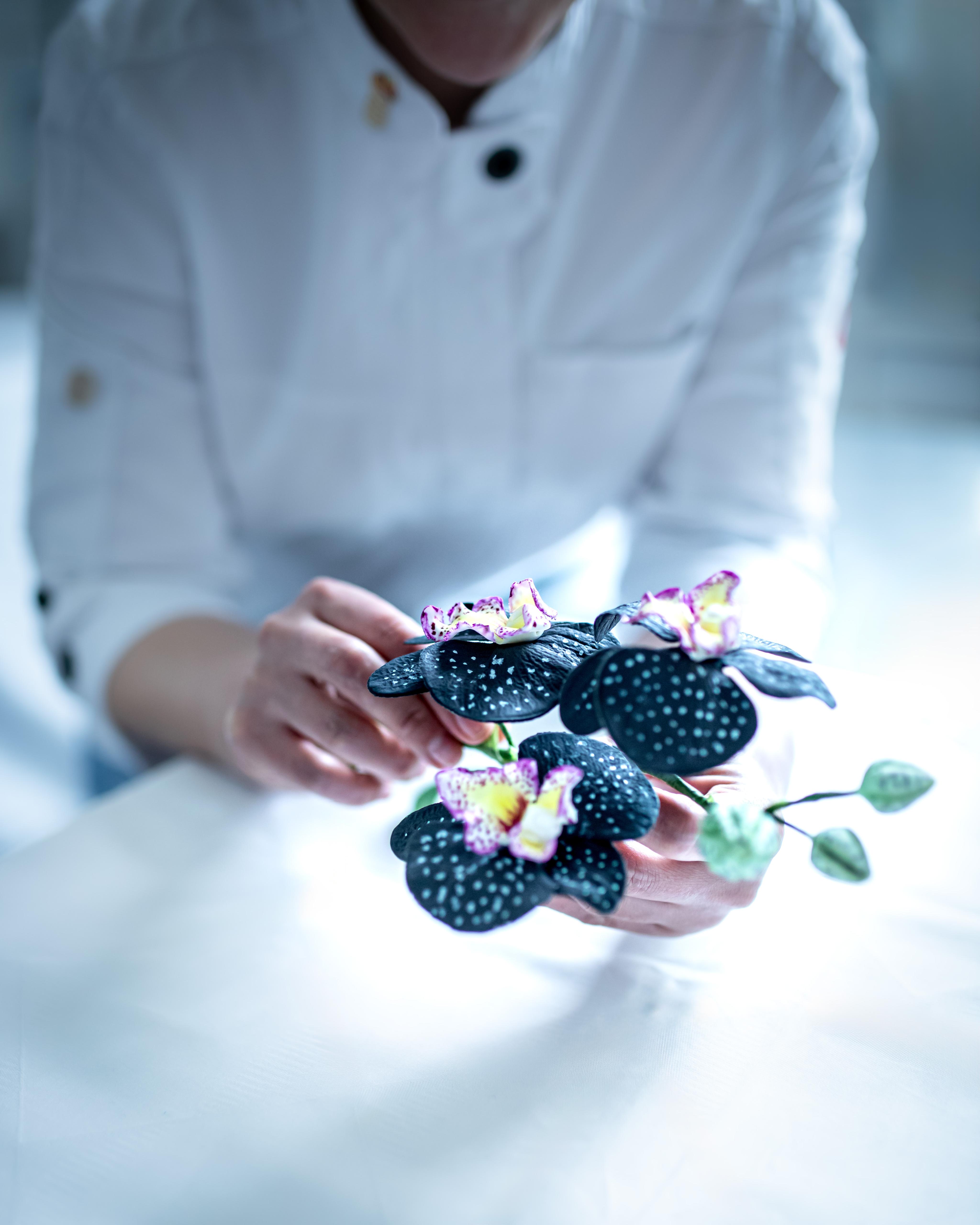 Konditorin in weißer Jacke hält handgefertigte schwarze Zucker-Orchideen mit filigranen Details – essbare Blütendekoration für Design-Torten aus Berlin