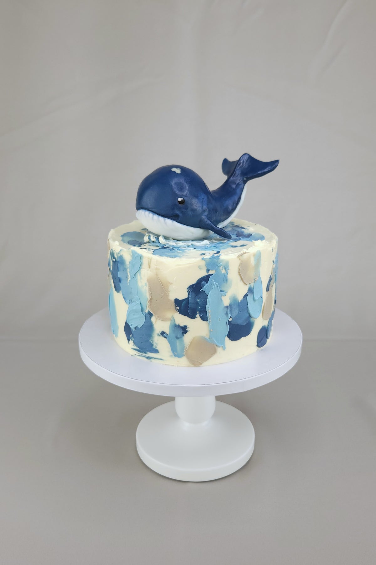Blau-cremefarbene Wal-Torte mit Wellendekoration und dunkelblauem Wal auf einem weißen Tortenständer