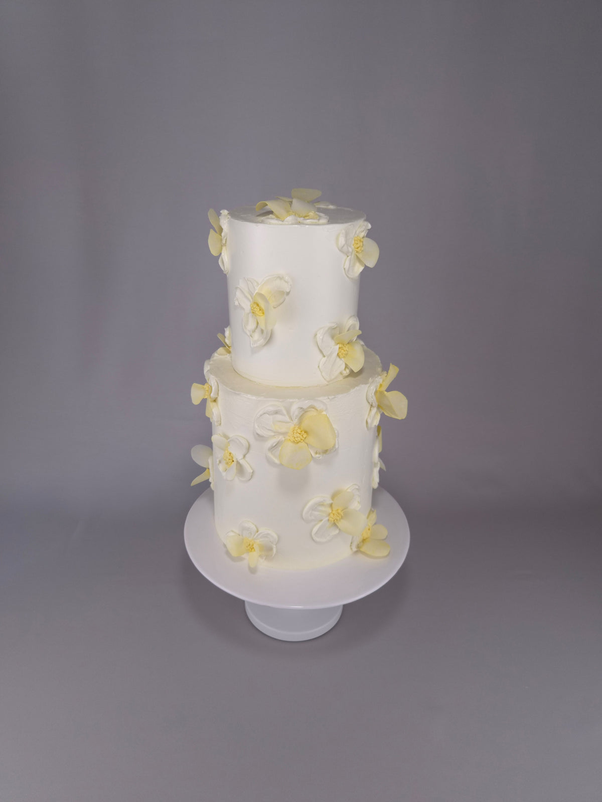 Zweistöckige weiße Hochzeitstorte mit weißen und hellgelben Blüten auf einem weißen Tortenständer