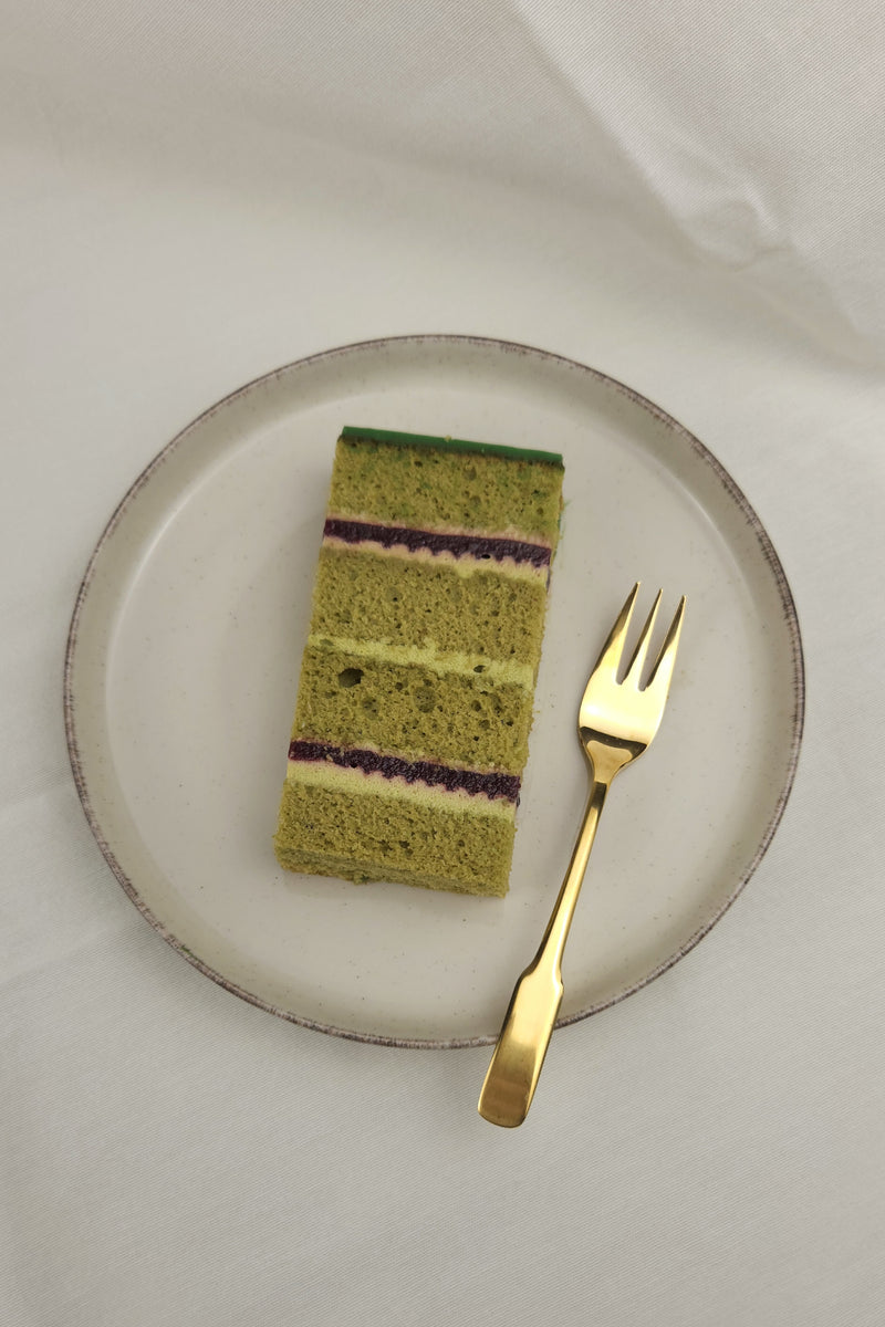 tück Matcha-Pistazien-Kirsch Torte auf Keramikteller mit goldener Kuchengabel – ein harmonisches Spiel aus grünem Tee, Nuss und Frucht